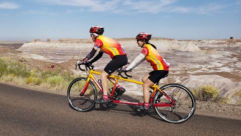 Two people on a yellow and red tandem bike ride past blue striped badlands.